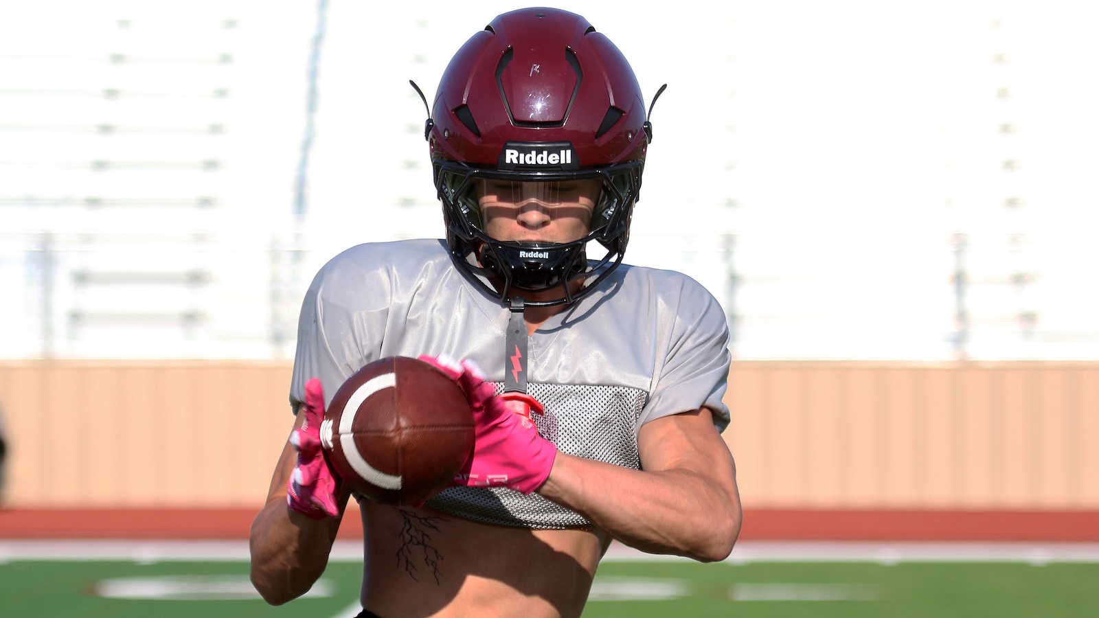American football player in a burgundy helmet and gray practice jersey catching a football on a sunny field, wearing pink gloves.