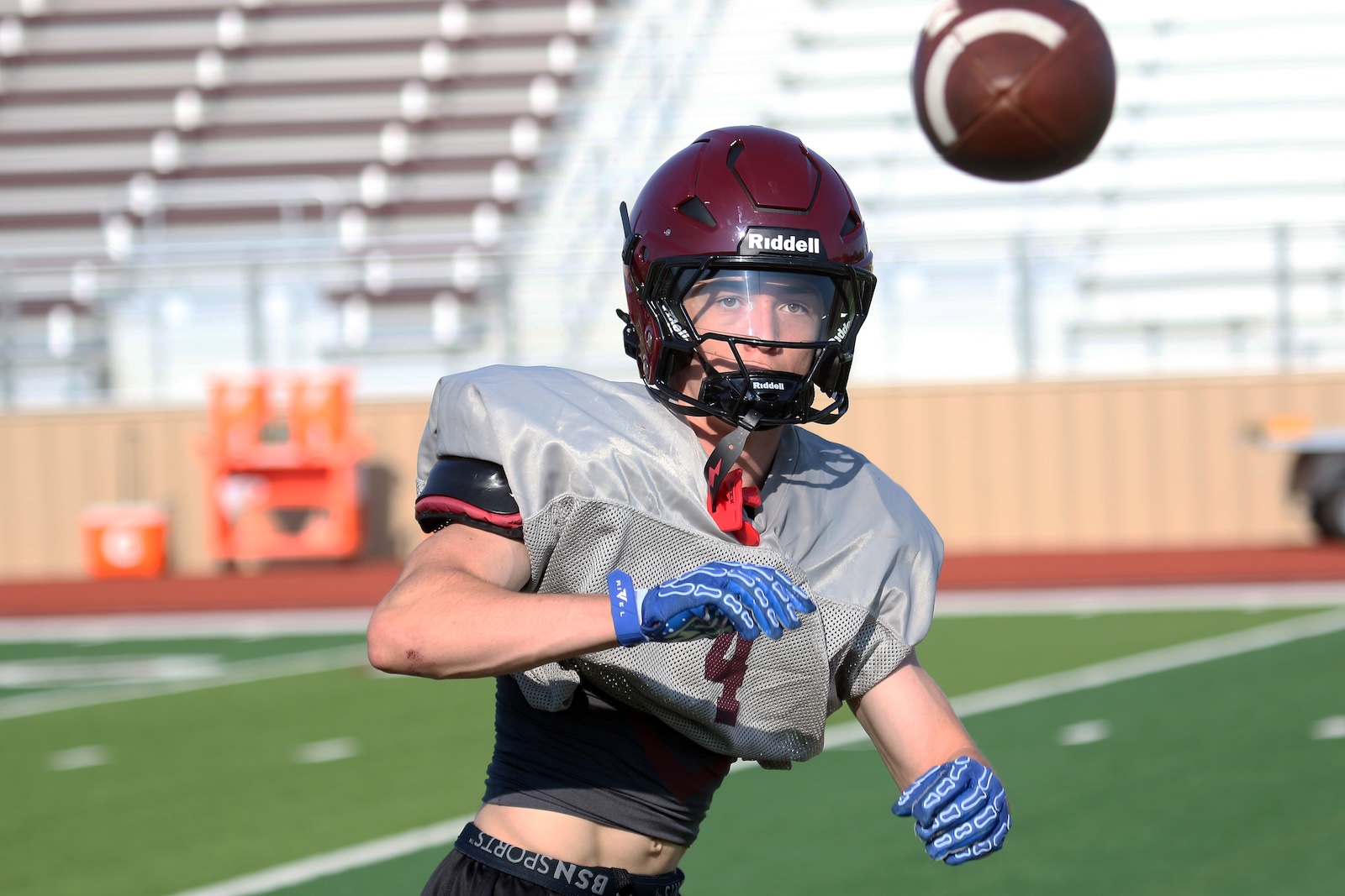 American football player in a maroon helmet reaching for a spiraling ball on a sunny practice field, wearing gray jersey and blue gloves