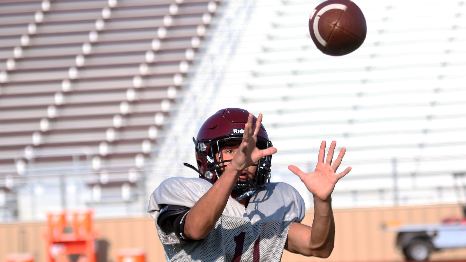 American football player in a maroon helmet reaches up to catch a football in midair during practice on a sunny day, bleachers in the background.