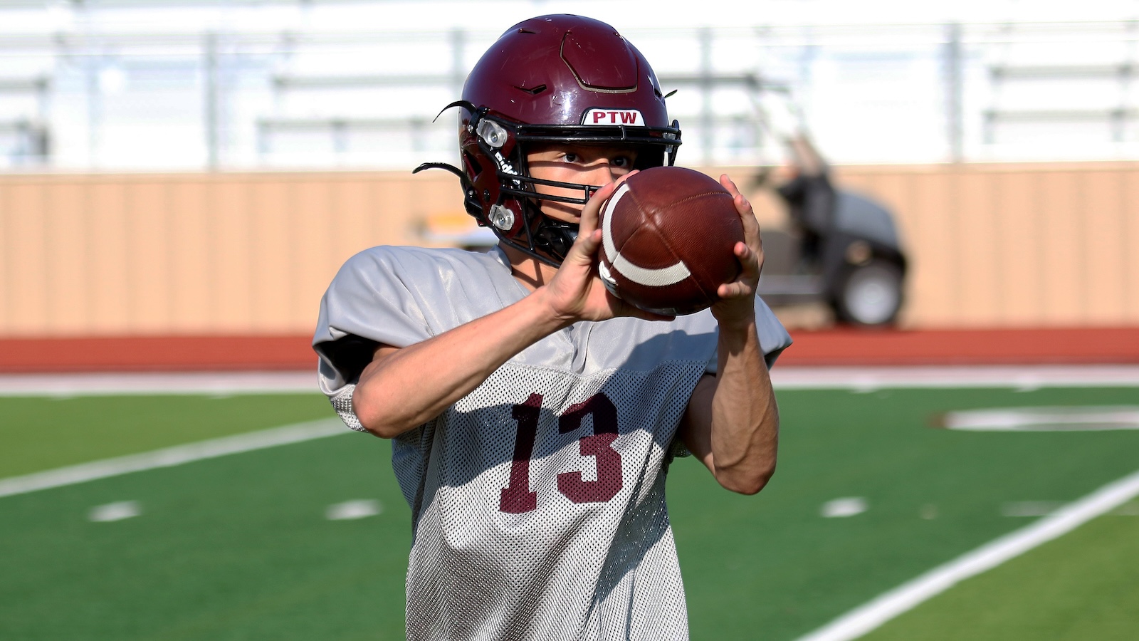 Football quarterback trainee in gray jersey (#13) catching a brown football with both hands on a turf field, helmet on wireless fence in background