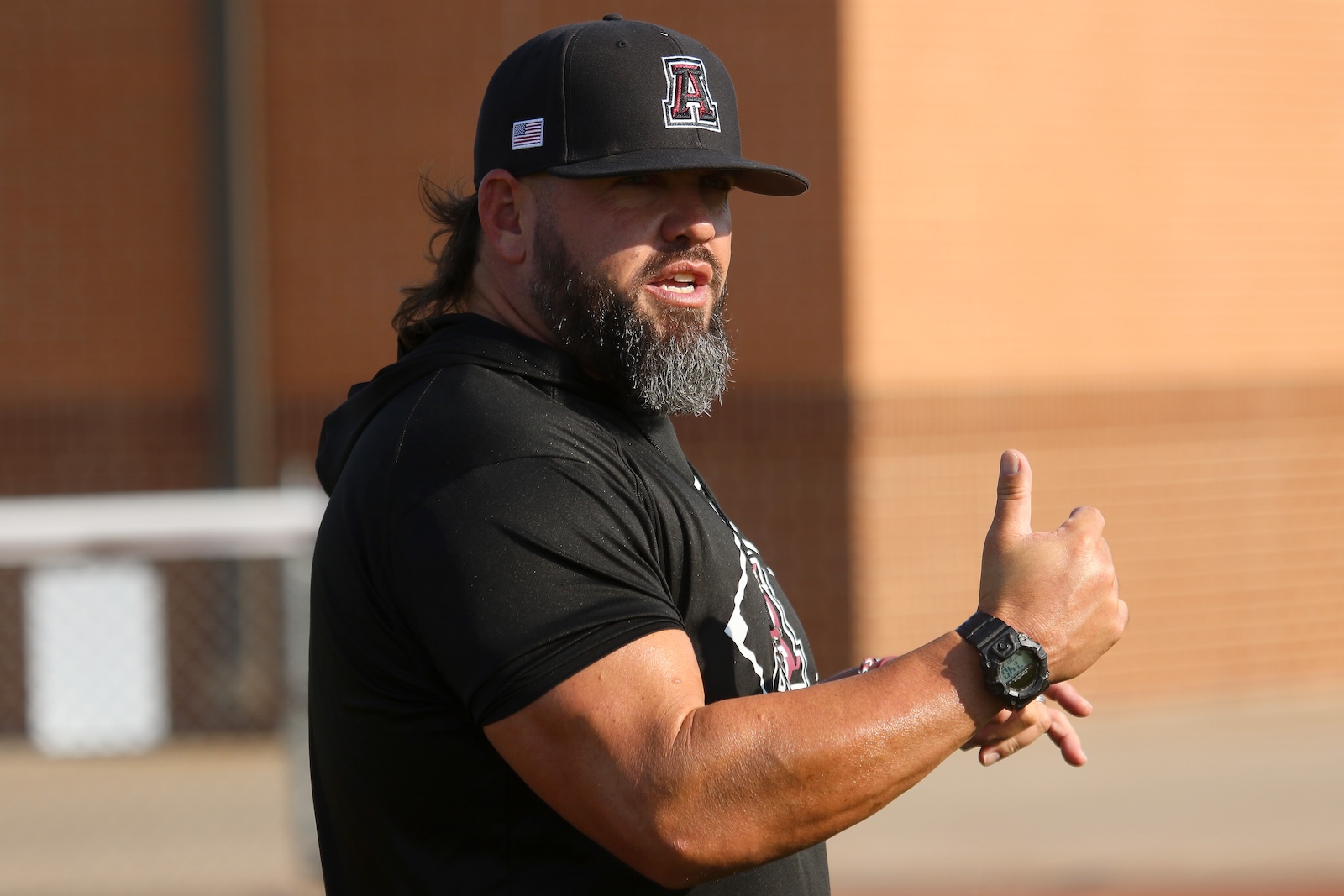 Man with a gray beard giving a thumbs-up, wearing a black cap with a patch and a black t-shirt outdoors by a brick wall