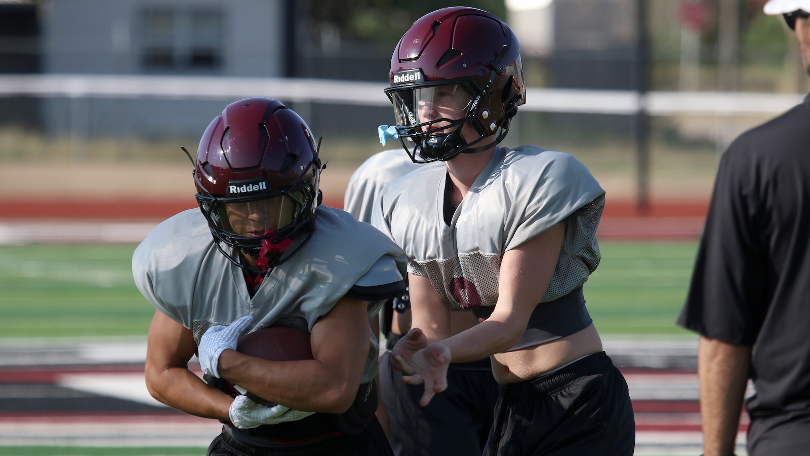 Two football players in gray practice jerseys and maroon helmets on a football field; one holds a ball close to his chest as they move.