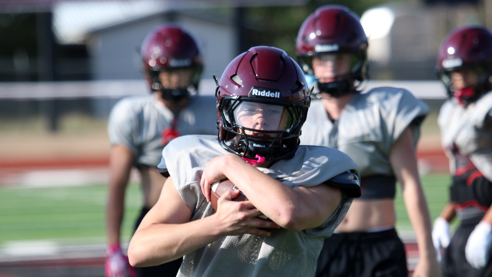 Football player in gray jersey and maroon helmet clutching a ball at the chest during practice, teammates blurred in the background