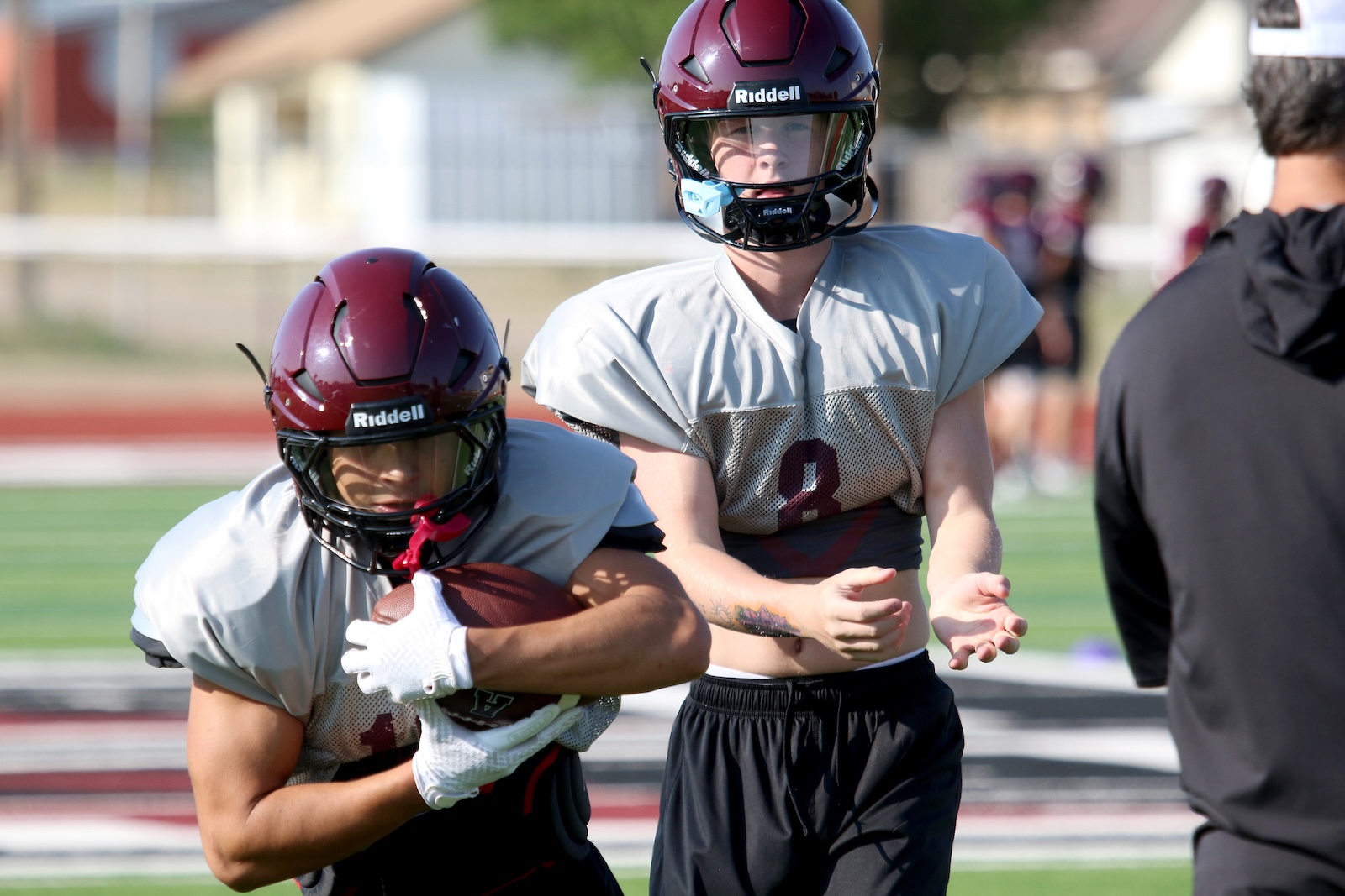 Two high school football players wearing maroon helmets and gray jerseys run on the field, one cradling the ball as a teammate helps block the way forward.