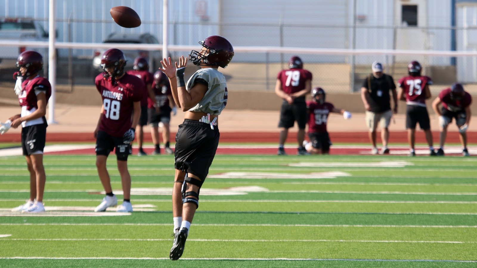Football player in a gray jersey catches a pass mid-air during practice on a green turf field, with teammates in maroon jerseys in the background.