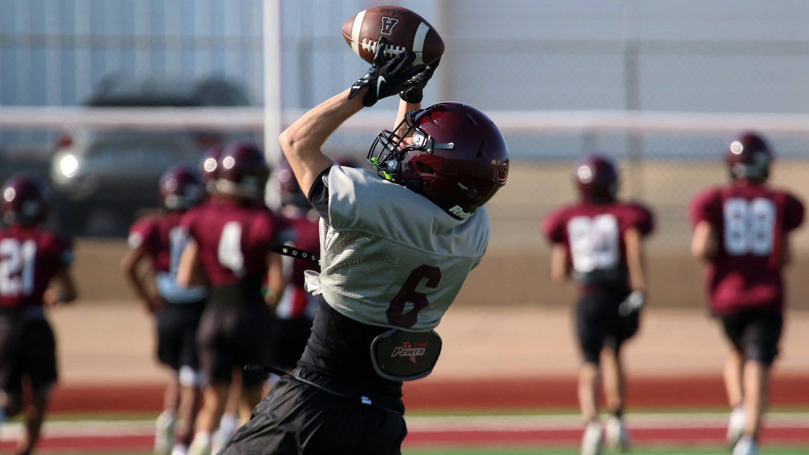 Football player in a gray jersey catches a ball overhead during practice, maroon helmets and teammates in the background