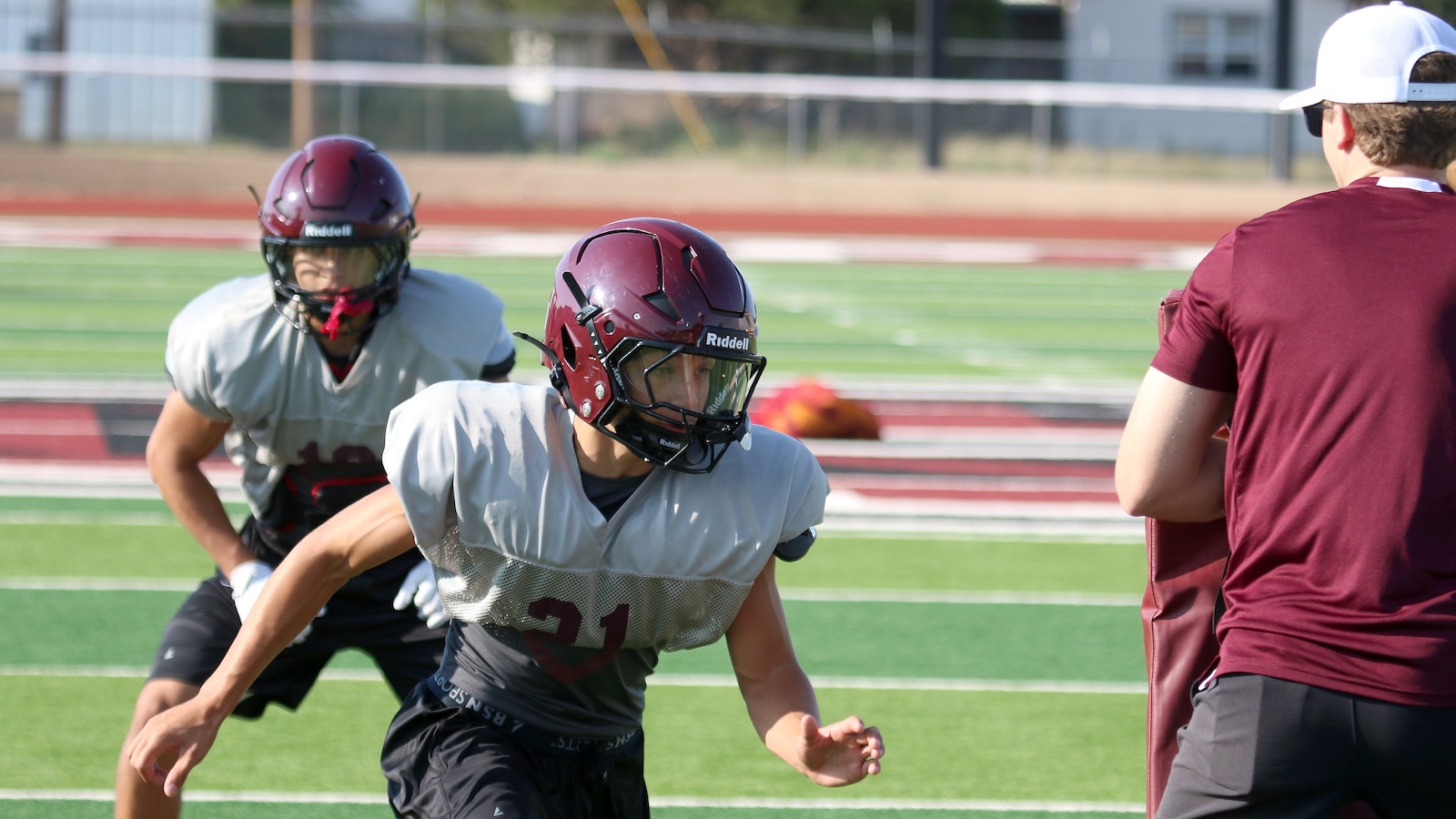 Two football players in gray practice jerseys sprint on a green turf while a coach in maroon watches on the right.
