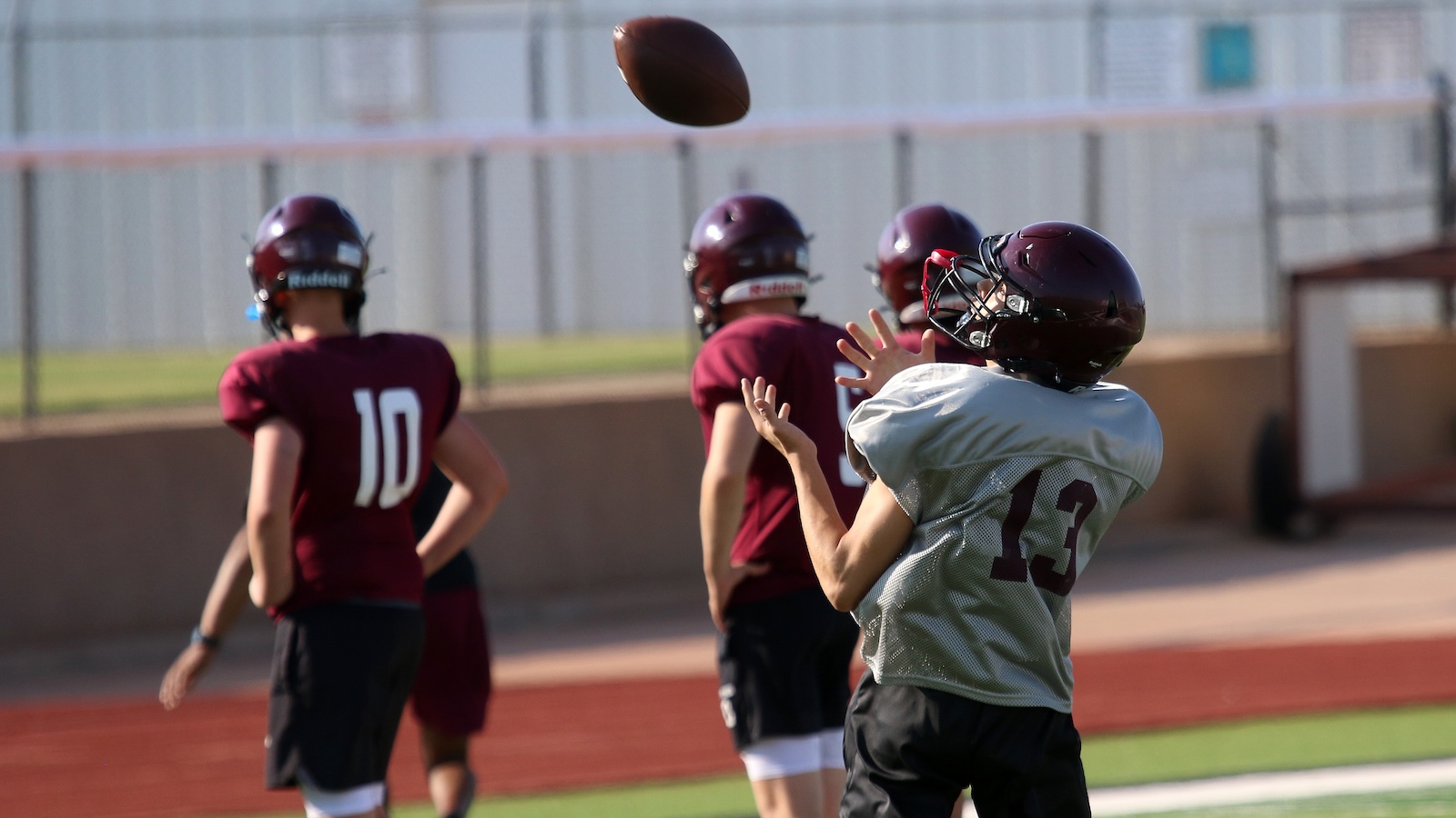 Youth football players practice on a field; a player in a gray jersey #13 reaches to catch a ball mid-air while others in maroon jerseys watch.