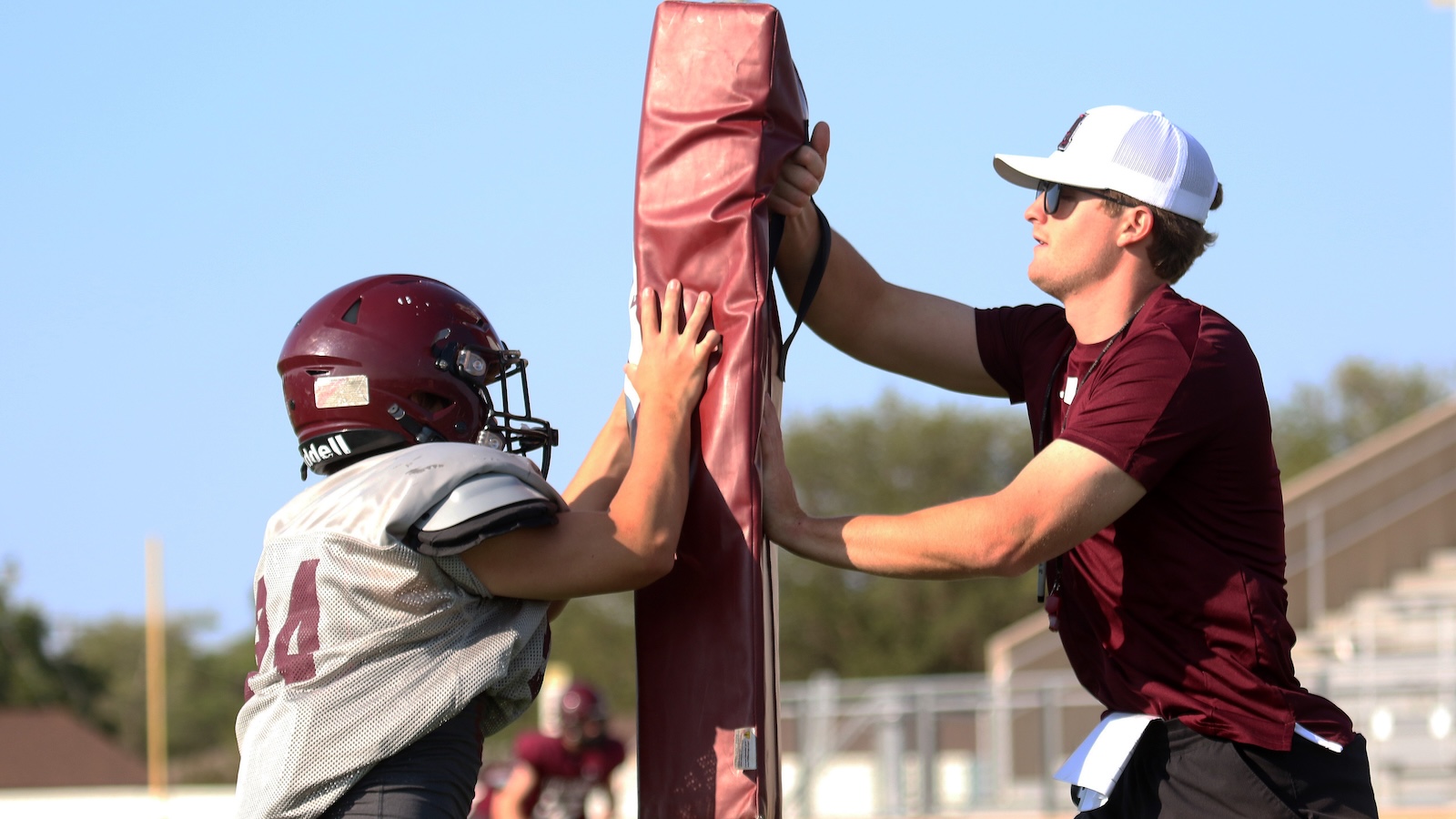 Two football players push against a padded blocking shield during practice, one in a maroon helmet and gray jersey, the other in a maroon shirt and white cap.