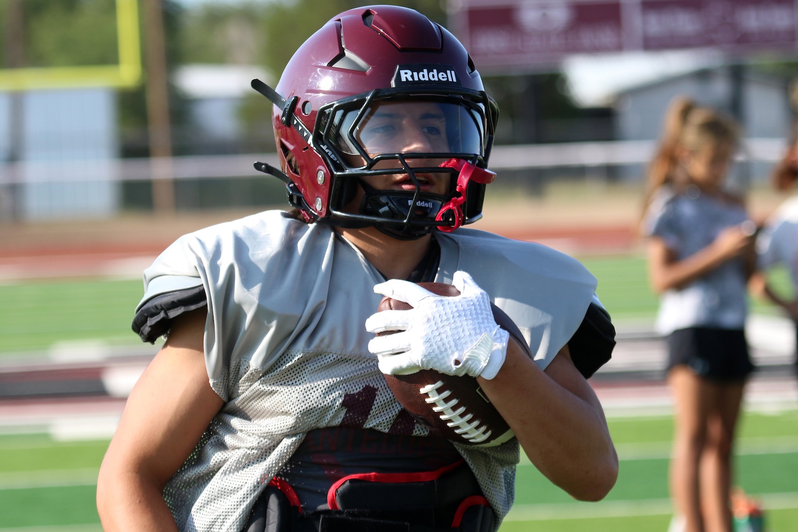 Football player in a maroon helmet running with ball on a turf field, wearing a gray jersey and white gloves.