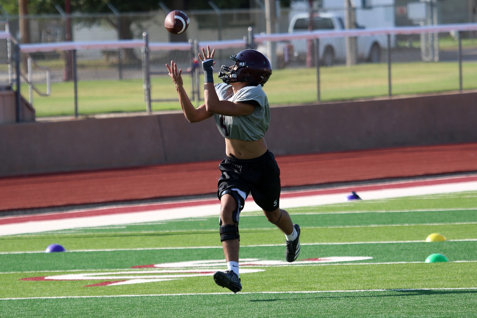 Youth football player in helmet and gray jersey reaches up to catch a ball on a green turf field during practice.