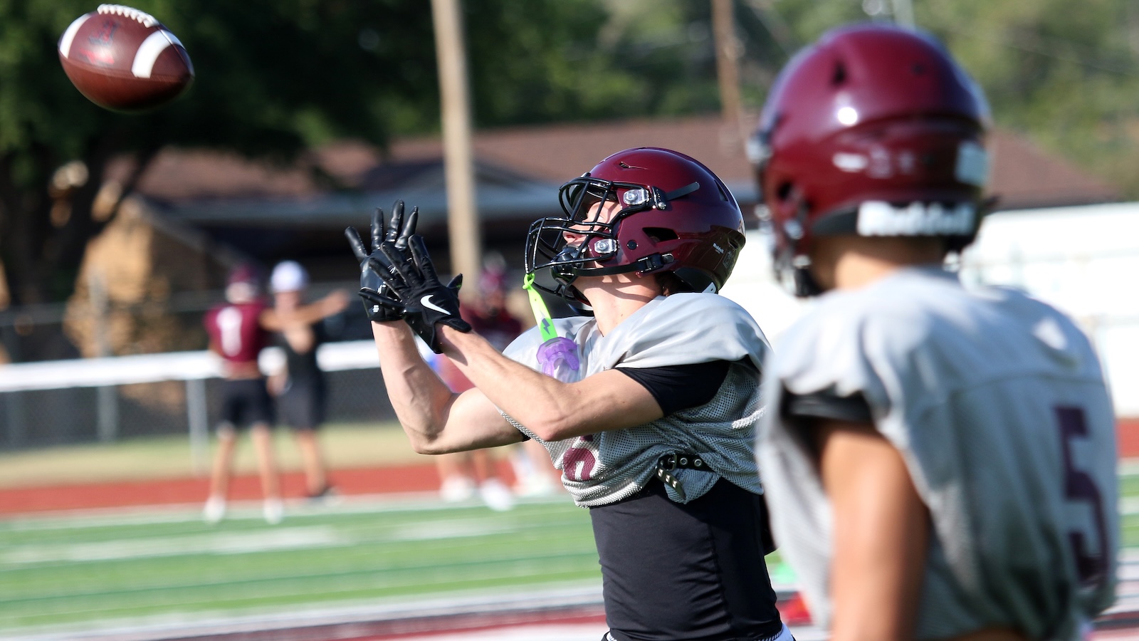 Football players in maroon helmets catch a high throw on a sunny practice field; one player stretches arms upward with gloves ready
