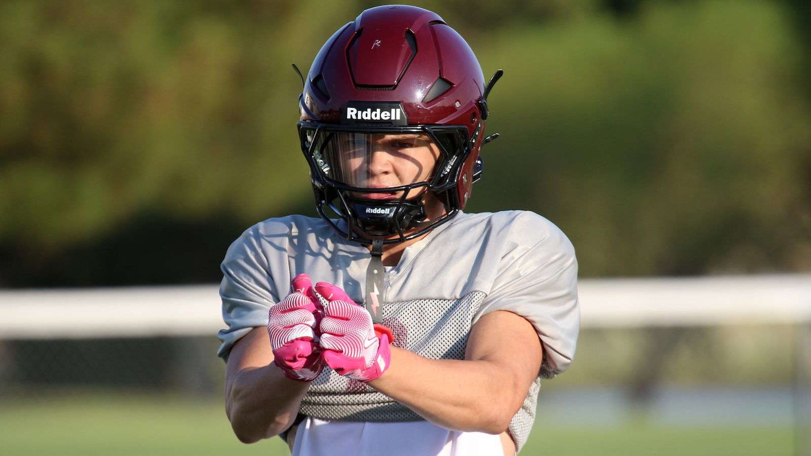 Youth football player in gray practice jersey and maroon helmet, wearing pink gloves, on a sunny field.