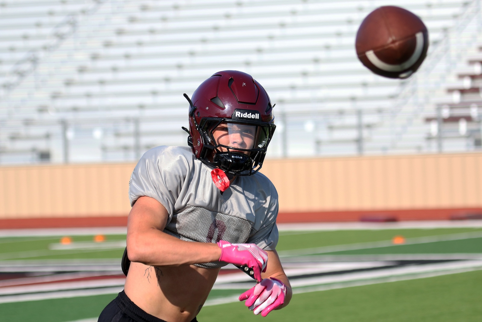 Female football player wearing maroon Riddell helmet and gray jersey, reaching for a football in the air on a practice field.