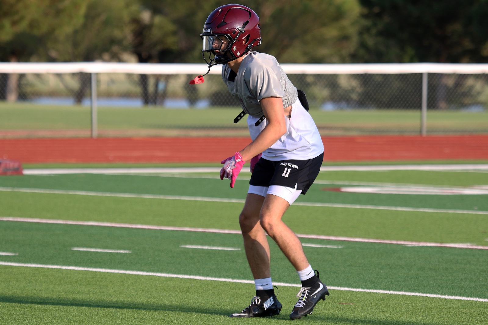 Football player in gray practice jersey and black shorts, maroon helmet and pink gloves, mid-sprint on a green artificial turf field.