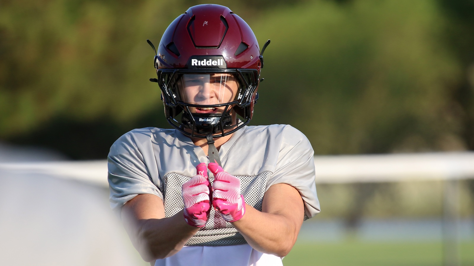Football player wearing a maroon Riddell helmet and pink gloves, adjusting chin strap on a practice field.
