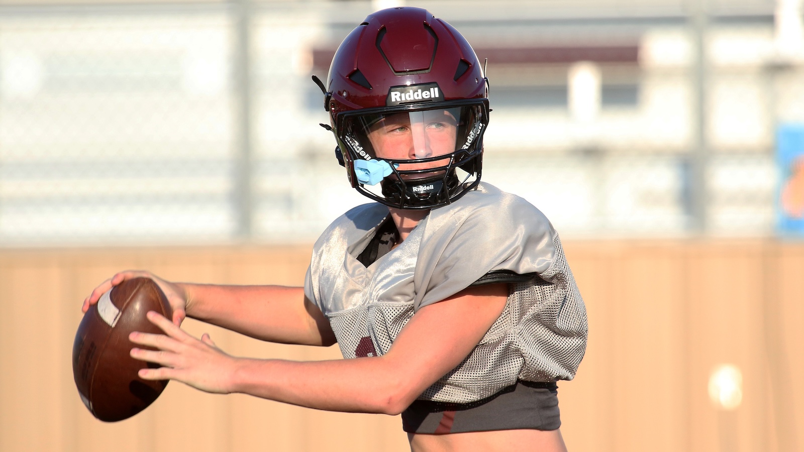 Quarterback in a maroon helmet throws a football during practice, eyes focused downfield in a gray jersey.