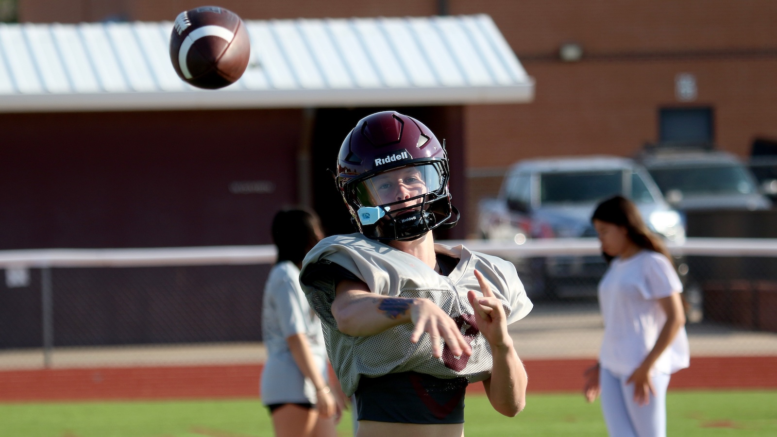 Football player in a maroon Riddell helmet and gray practice jersey mid-throw on a sports field, with teammates and a building in the background.