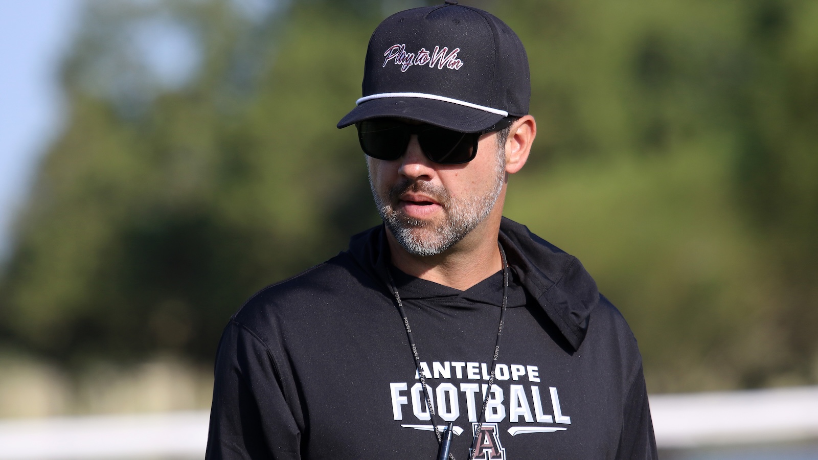 Man wearing a black cap with a 'Play to Win' logo and dark sunglasses, outdoors.