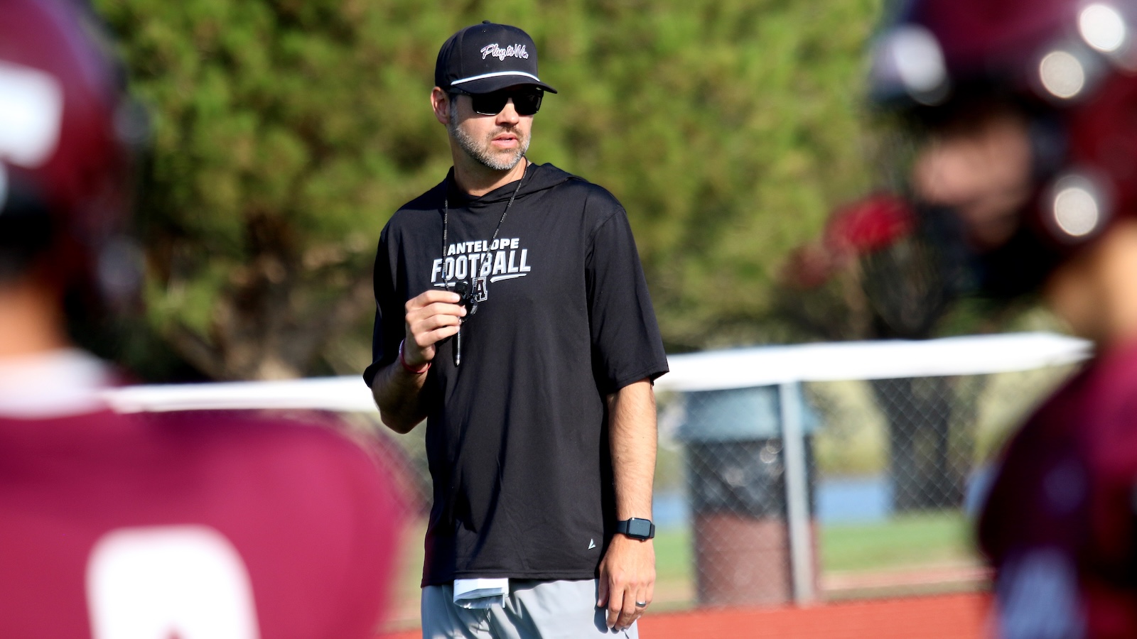 Male football coach in a black shirt and cap stands on a practice field, holding a whistle and wearing sunglasses.