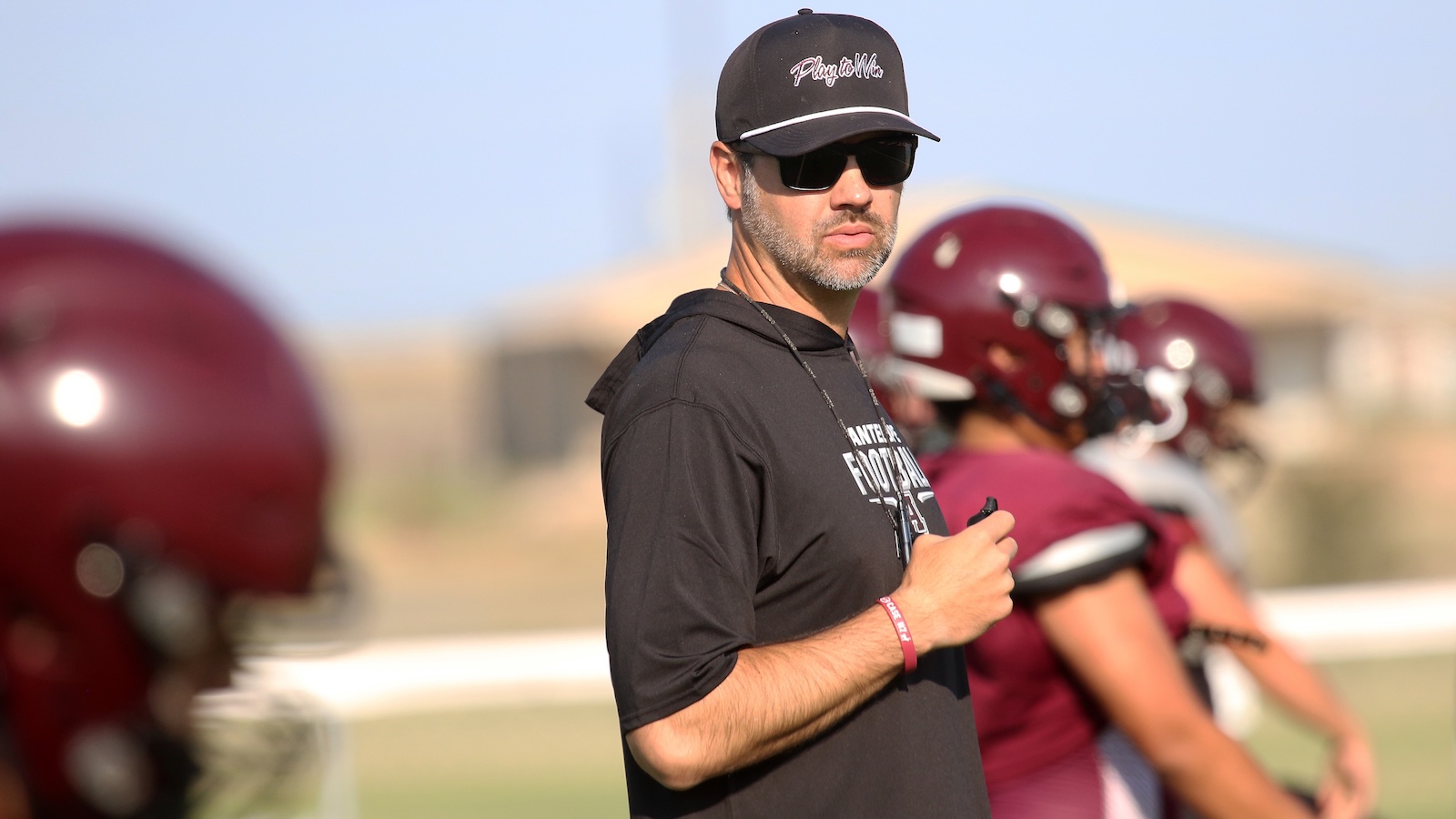 Coach wearing a black cap, sunglasses, and a lanyard watches football practice on a sunny day, with players in maroon helmets lined up in the background.
