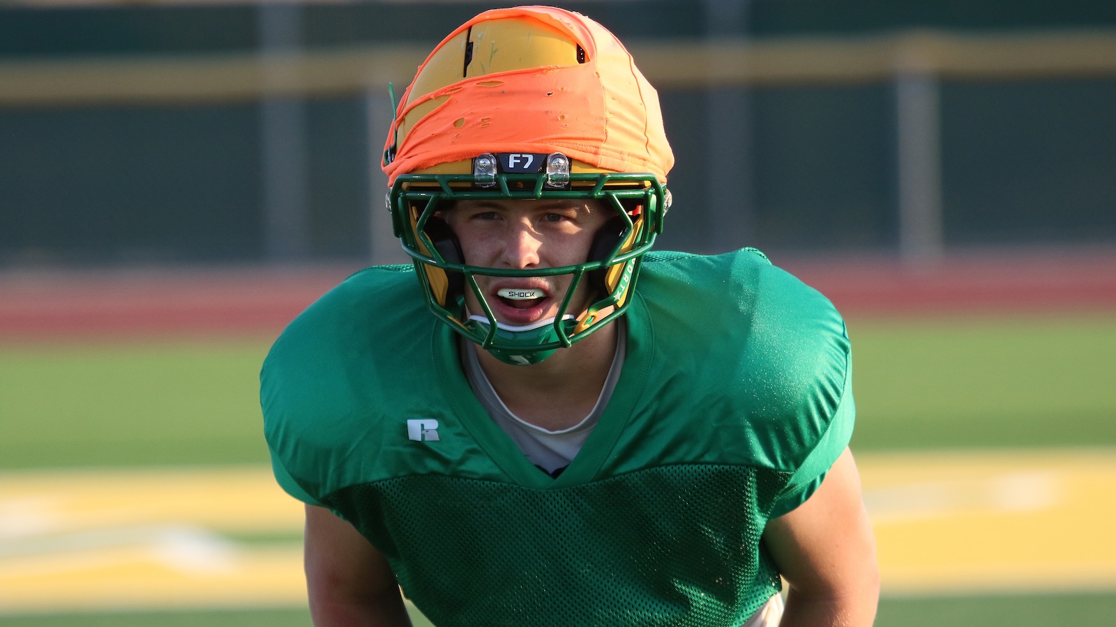 Football player in a green jersey crouches forward on a football field, wearing an orange helmet and mouthguard.