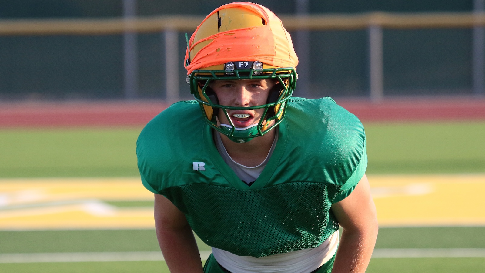 Football player in a green practice jersey crouches on a field, wearing an orange helmet with a green facemask.