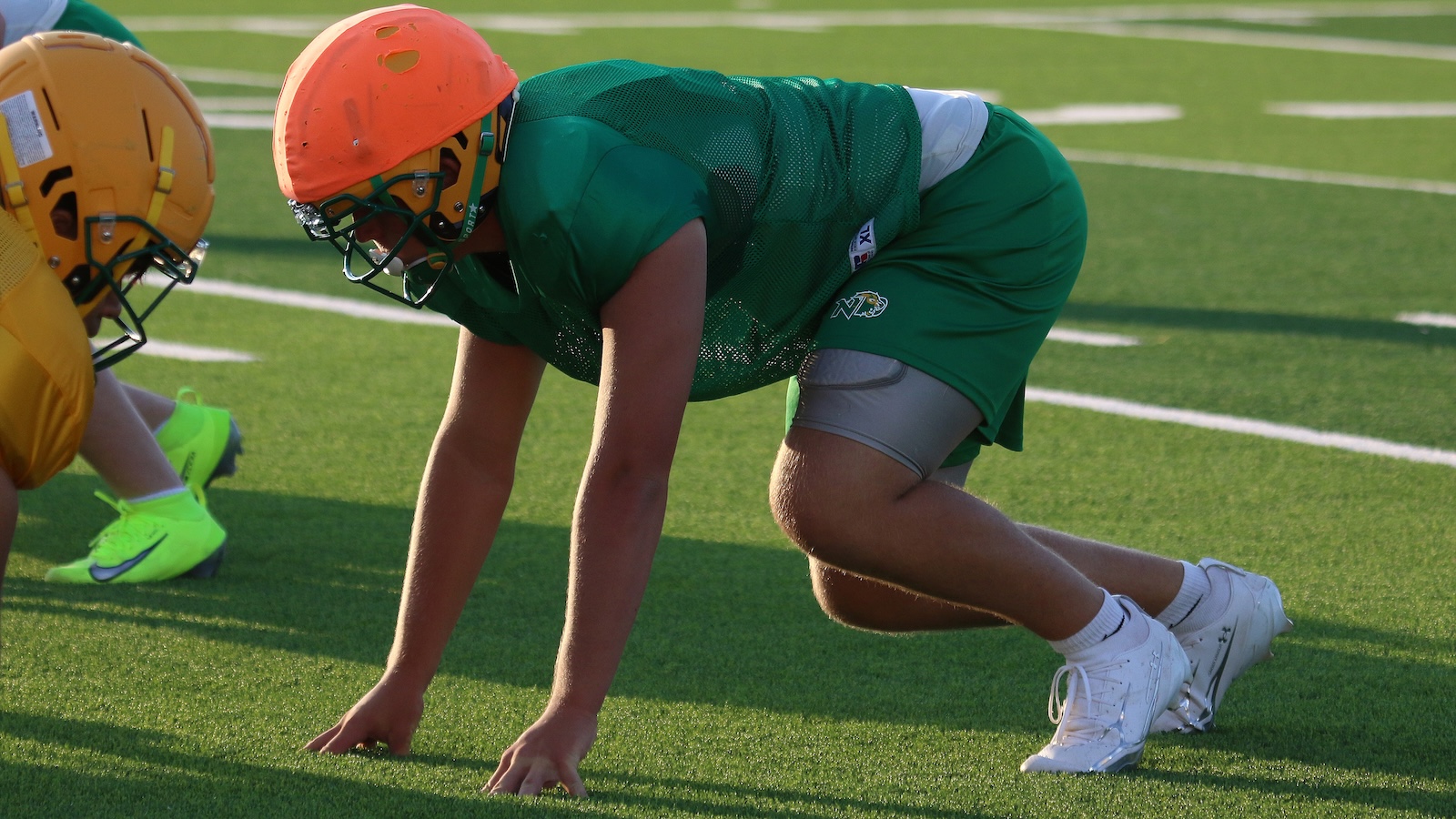 Two football linemen at the line of scrimmage: a green-jersey player with an orange helmet facing a yellow-uniform opponent on a grass field.