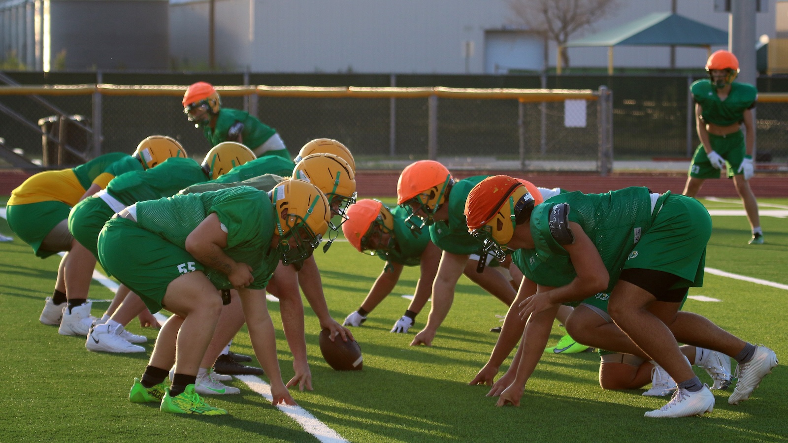 Football players in green uniforms line up at the line of scrimmage on a sunny practice field, with centers preparing to snap the ball.