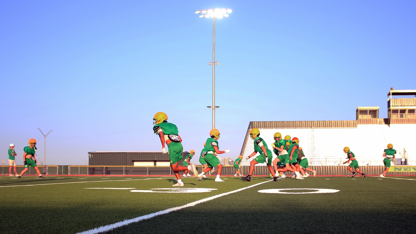 Youth football players in green uniforms running on a field during practice, with bleachers and a tall floodlight behind them.