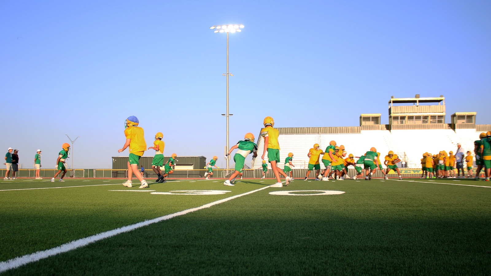 Group of young football players in yellow and green uniforms practicing on a sunny football field, with bleachers in the background.