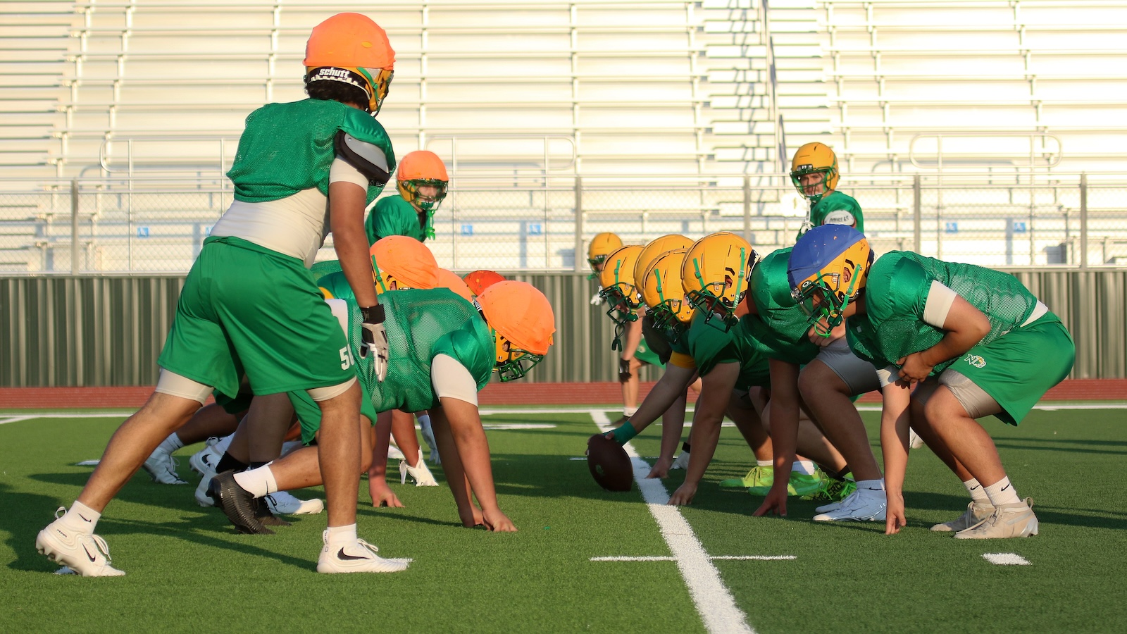 American football players in green uniforms crouch at the line of scrimmage on a turf field, wearing orange helmets and ready to snap the ball.