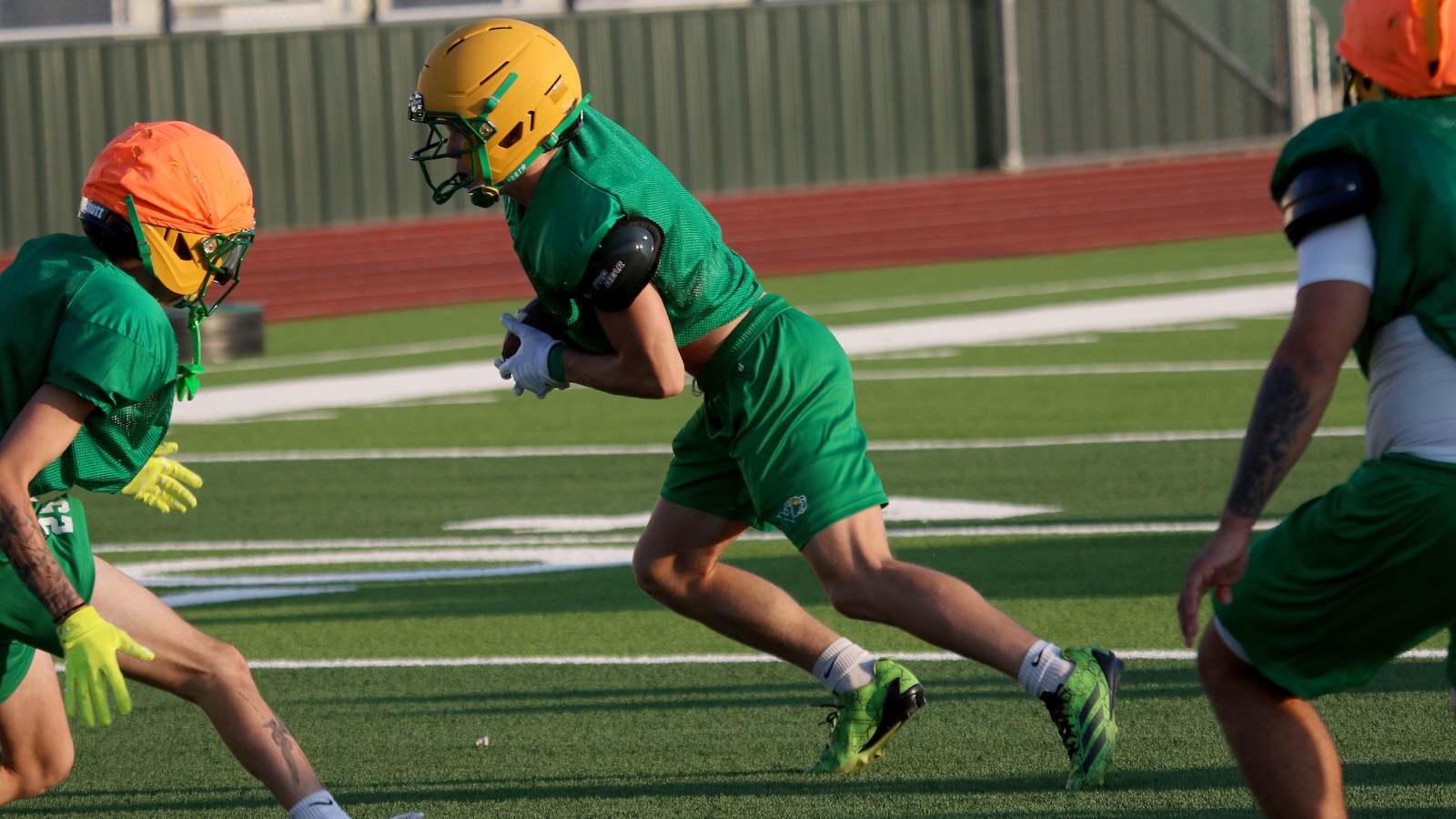 Green-uniform football players sprint on a practice field; central player carries the ball and is tackled by teammates in orange helmets.