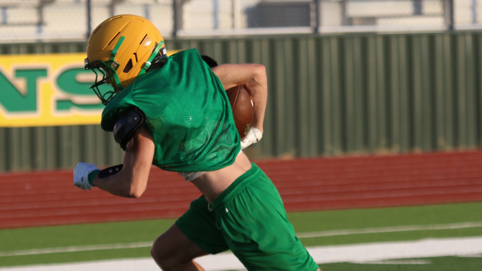 American football player wearing a yellow helmet and green jersey/shorts runs with the ball on a field during a game.