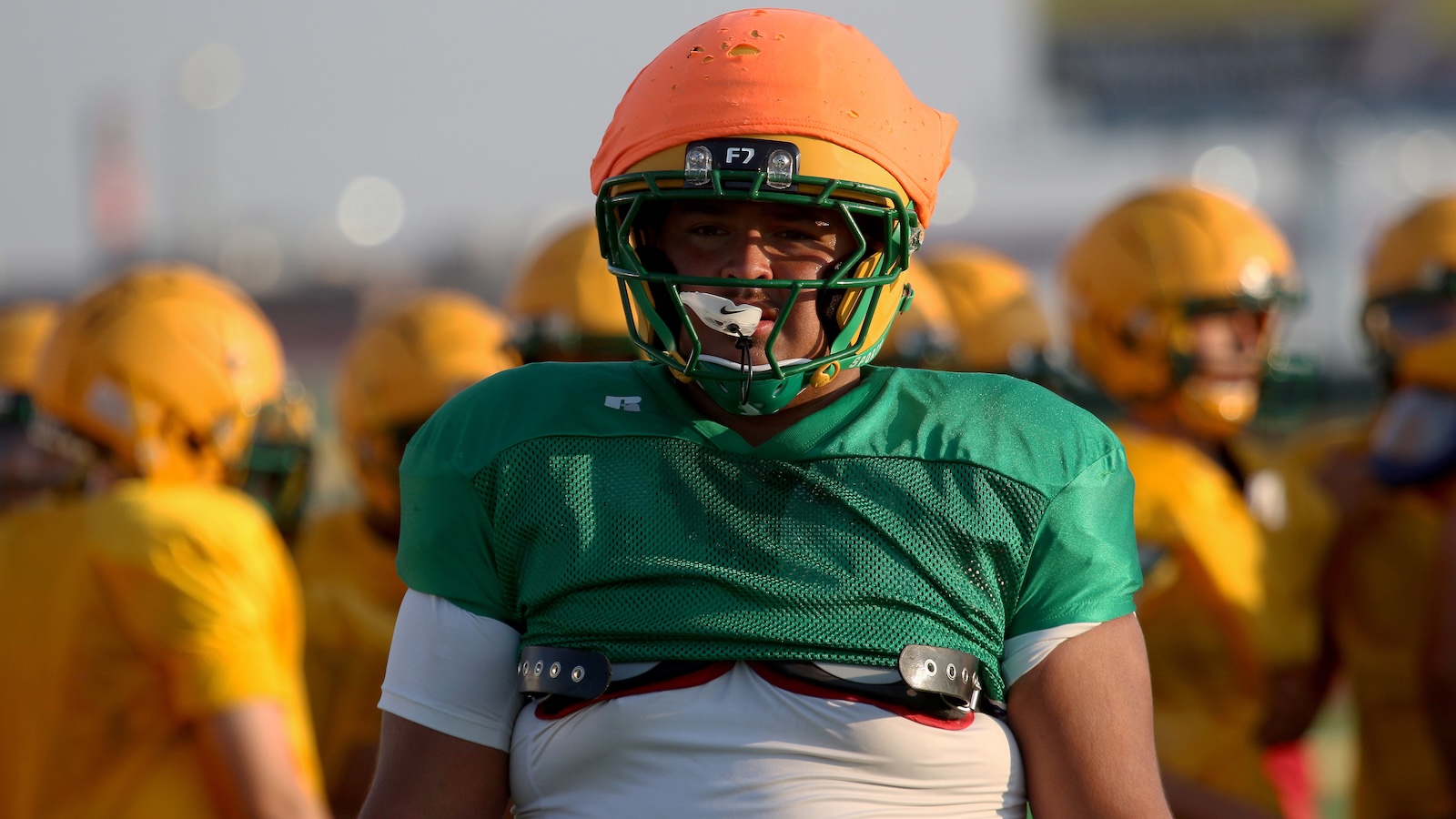Football player in a green practice jersey and orange helmet with mouthguard, surrounded by teammates in yellow uniforms during practice.