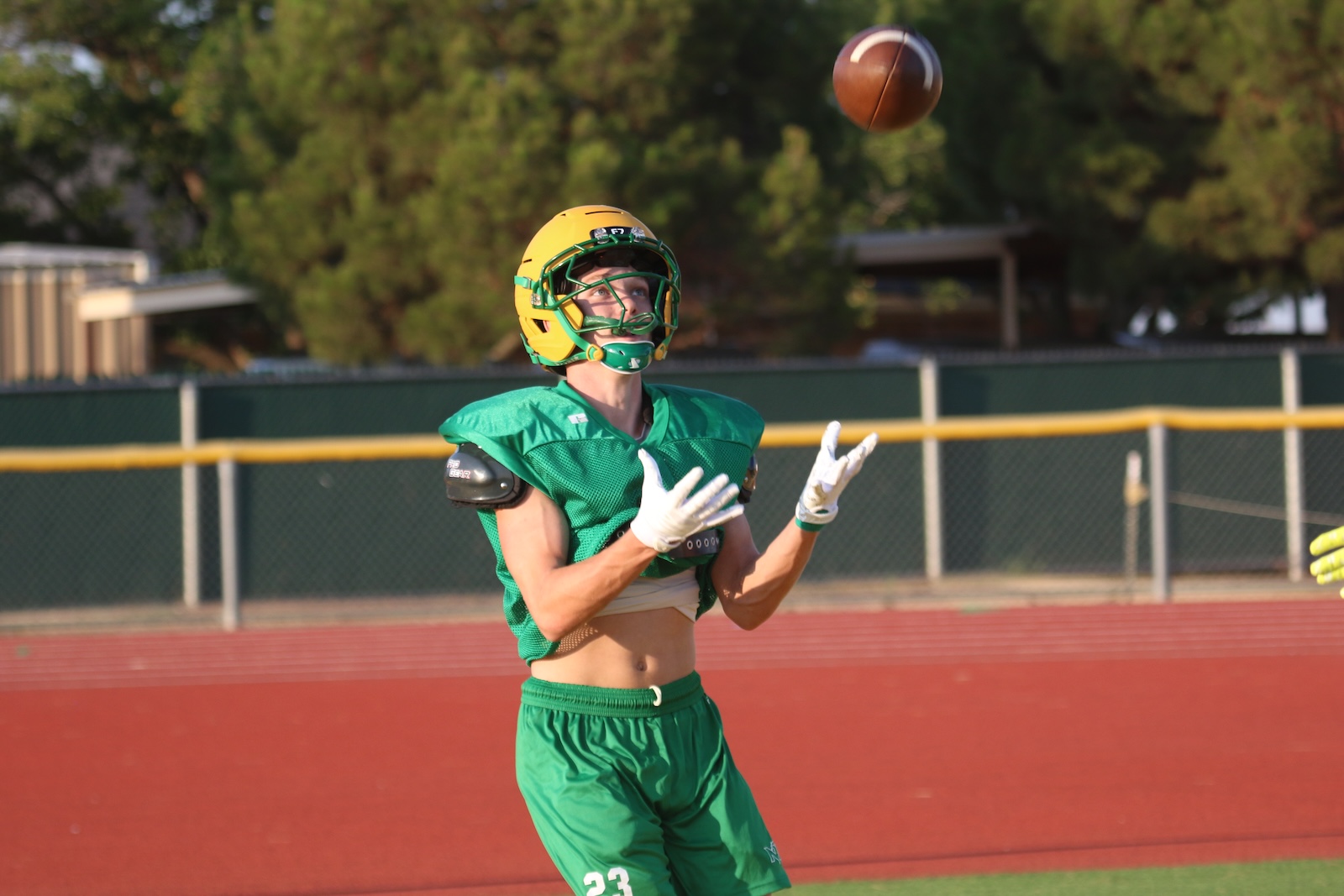 Football player in green uniform and yellow helmet watches a ball descend toward hands, on a track field with a chain-link fence in the background.