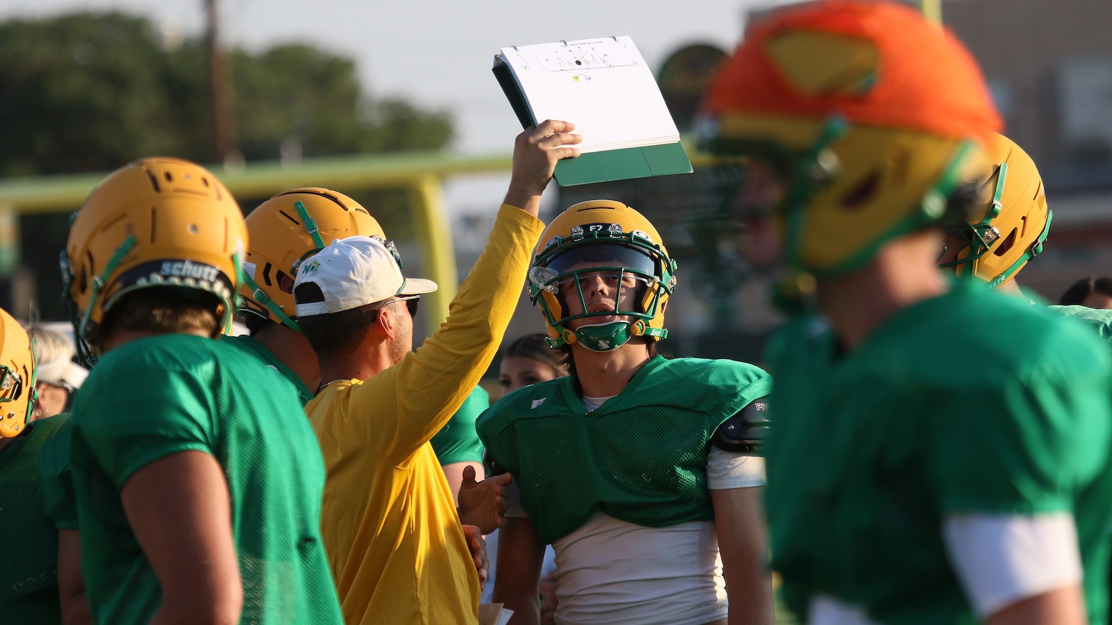 Football coach raises a clipboard to a huddled team during practice on a sunny day.
