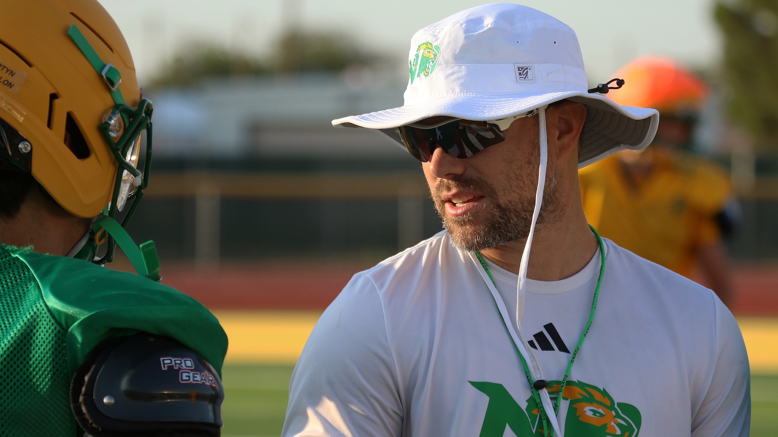 Coach with white bucket hat and sunglasses talks to a football player in green gear on the field at practice.