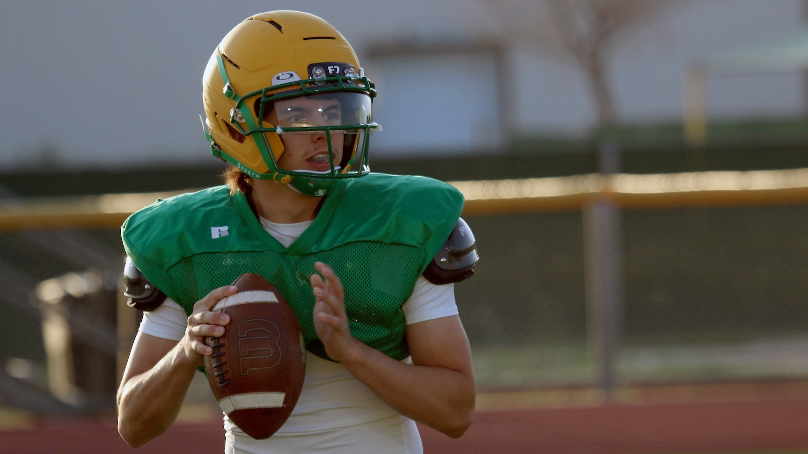 Football quarterback in a green practice jersey and yellow helmet holding a ball, looking to pass on a track field.