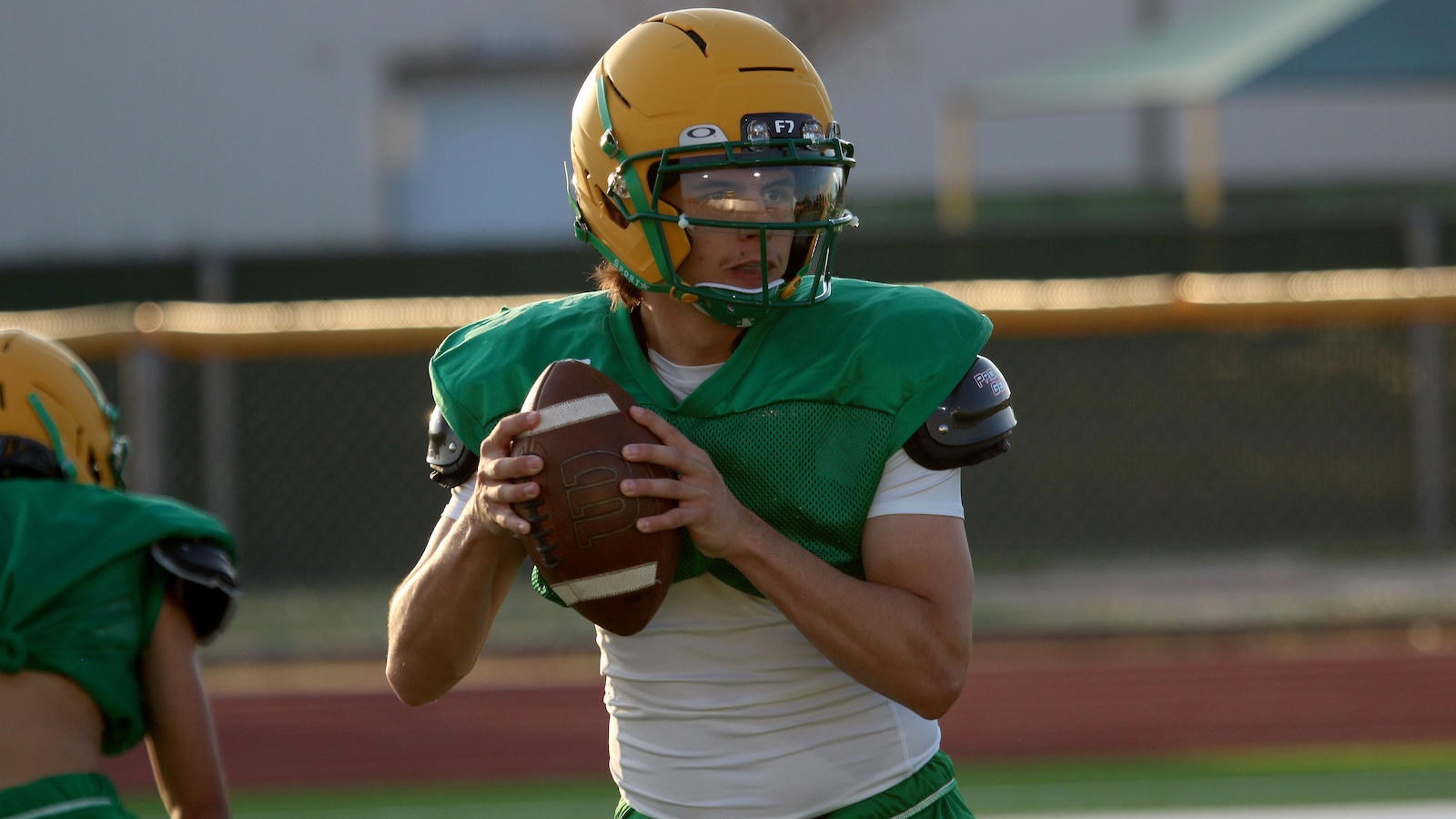 Young football quarterback in a green practice jersey and yellow helmet prepares to throw a ball on the field.