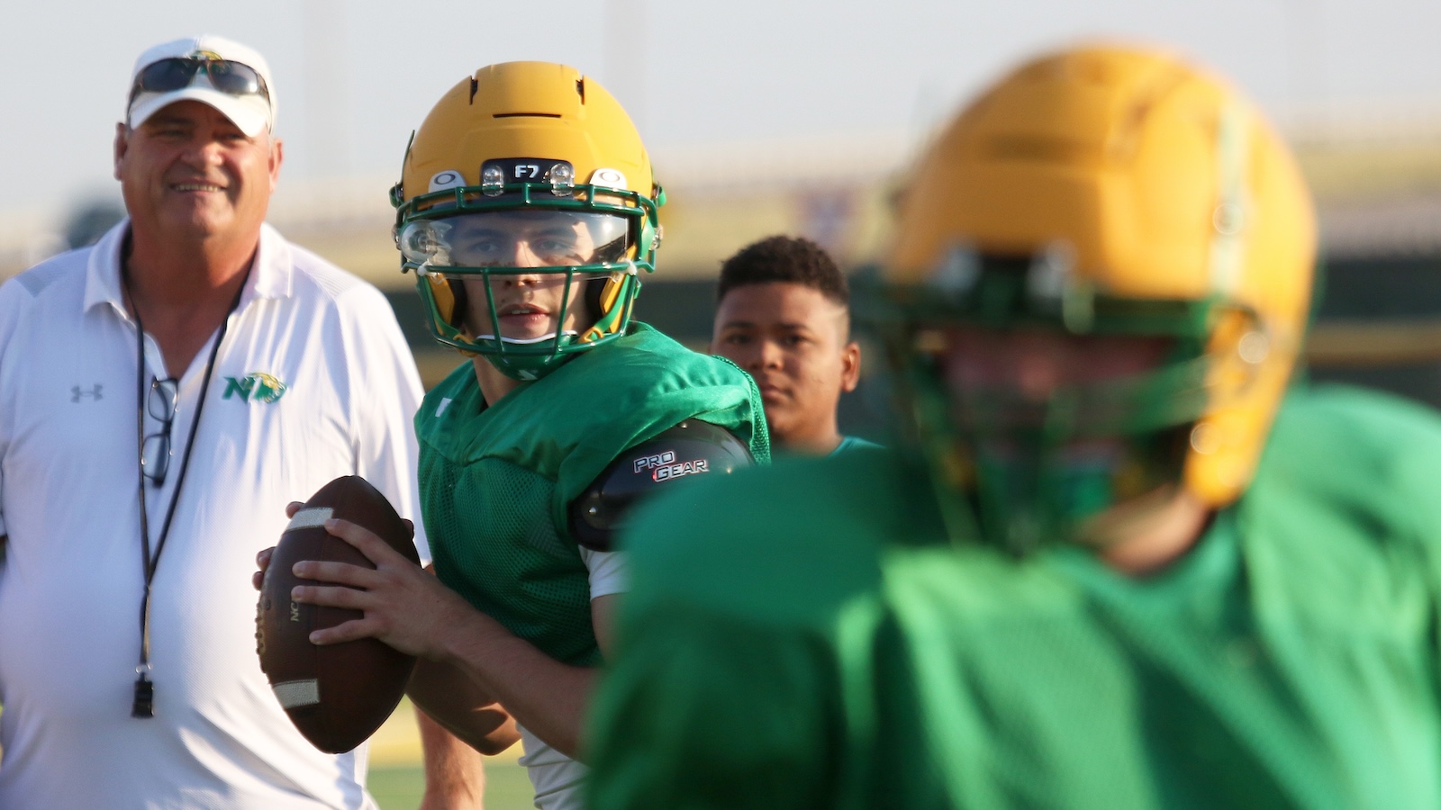 Quarterback in green jersey and yellow helmet holds a football while coach watches from the left; other players in green gear in background.