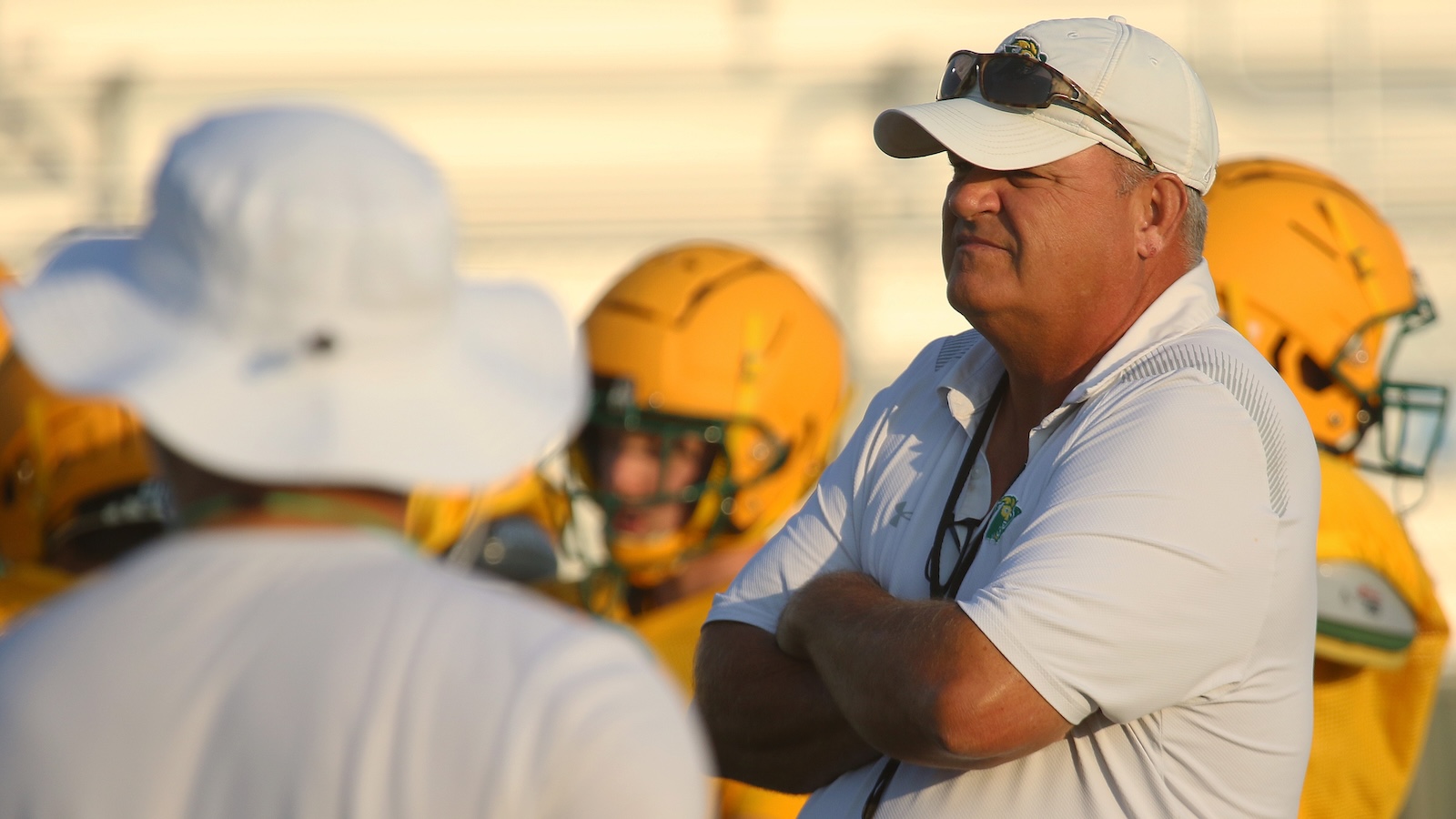 Coach in white polo and cap with sunglasses stands with arms crossed while yellow-helmeted players listen on a football field, sunlit.