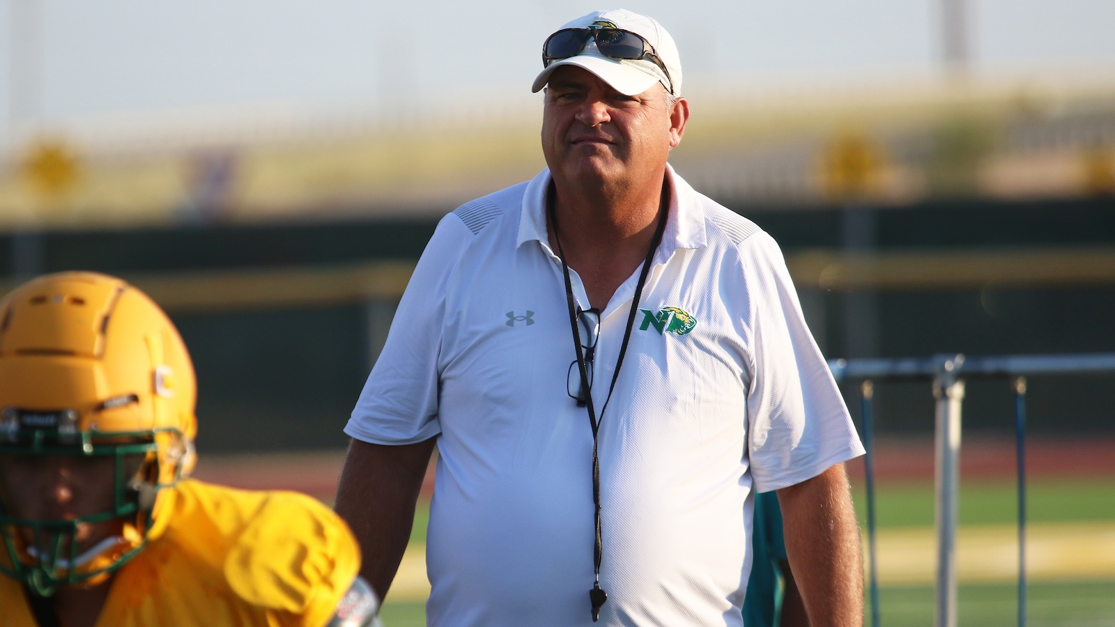 Football coach wearing a white polo and cap walks beside a player in a yellow helmet on the field.