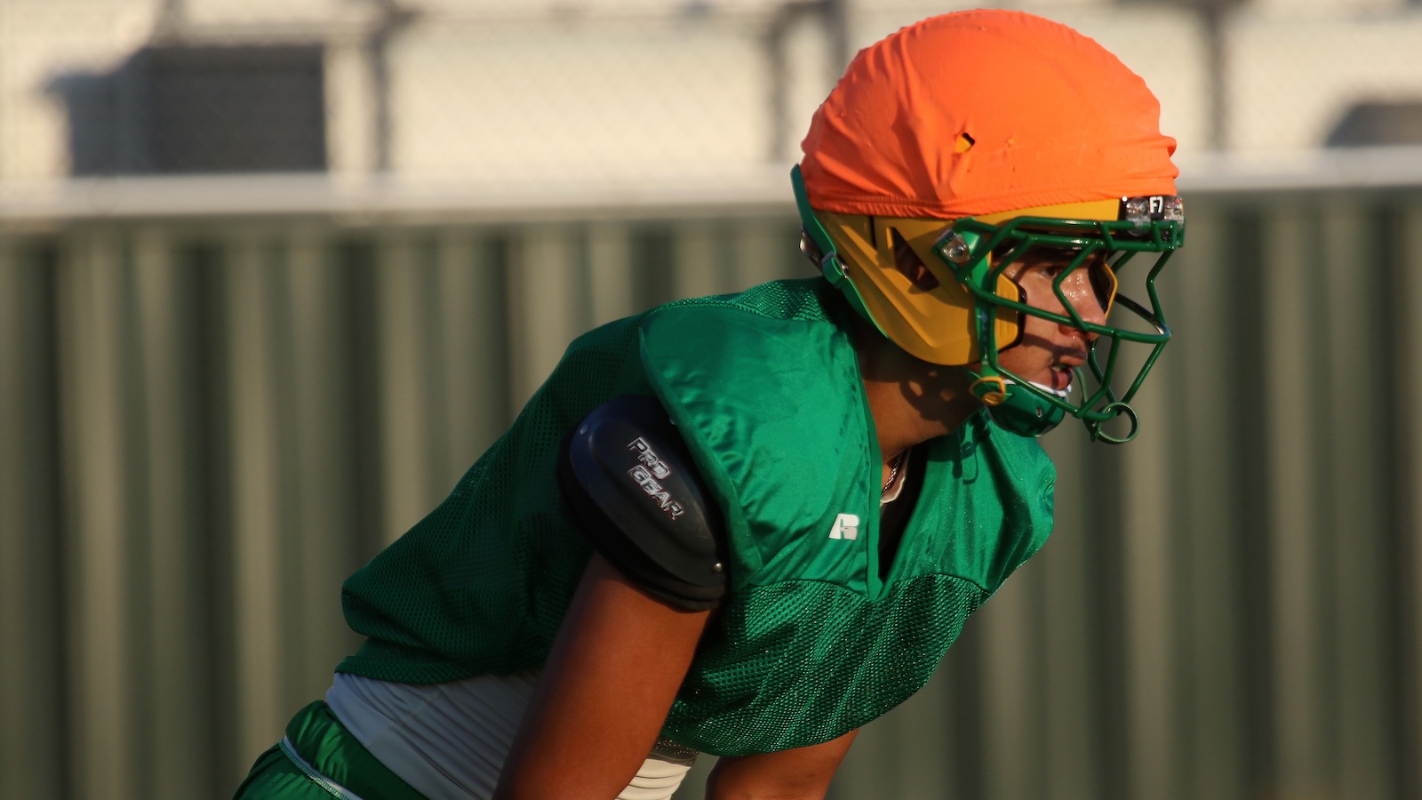 Young football player in green uniform and orange helmet crouches in a ready stance on a field behind a metal fence route.