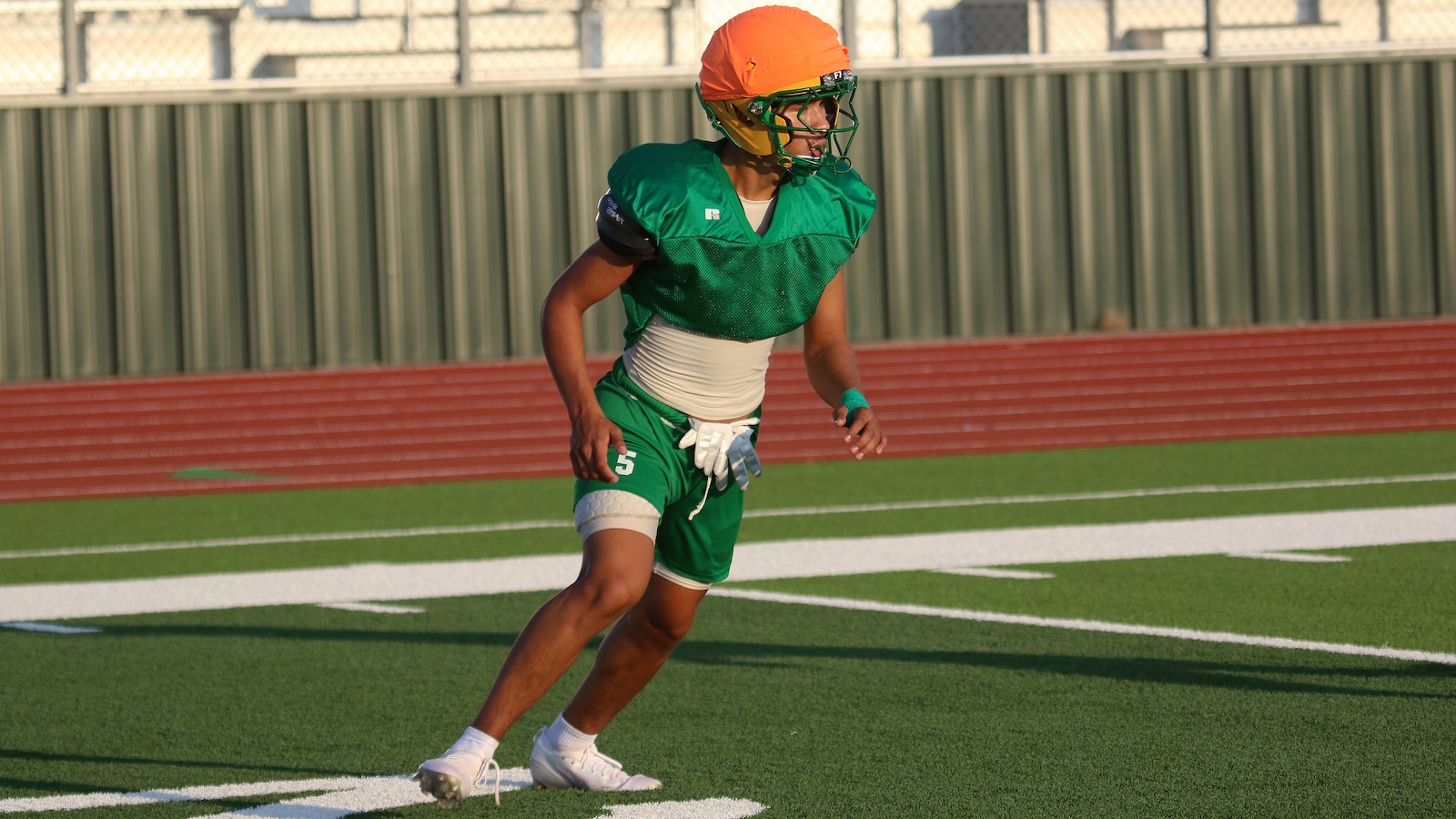 Football player in a green jersey and orange helmet sprinting on artificial turf field.