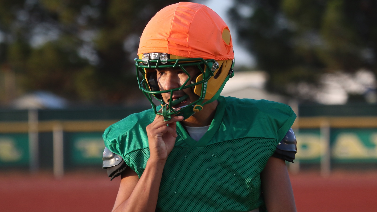 Football player in an orange helmet and green jersey adjusts his facemask on the field.