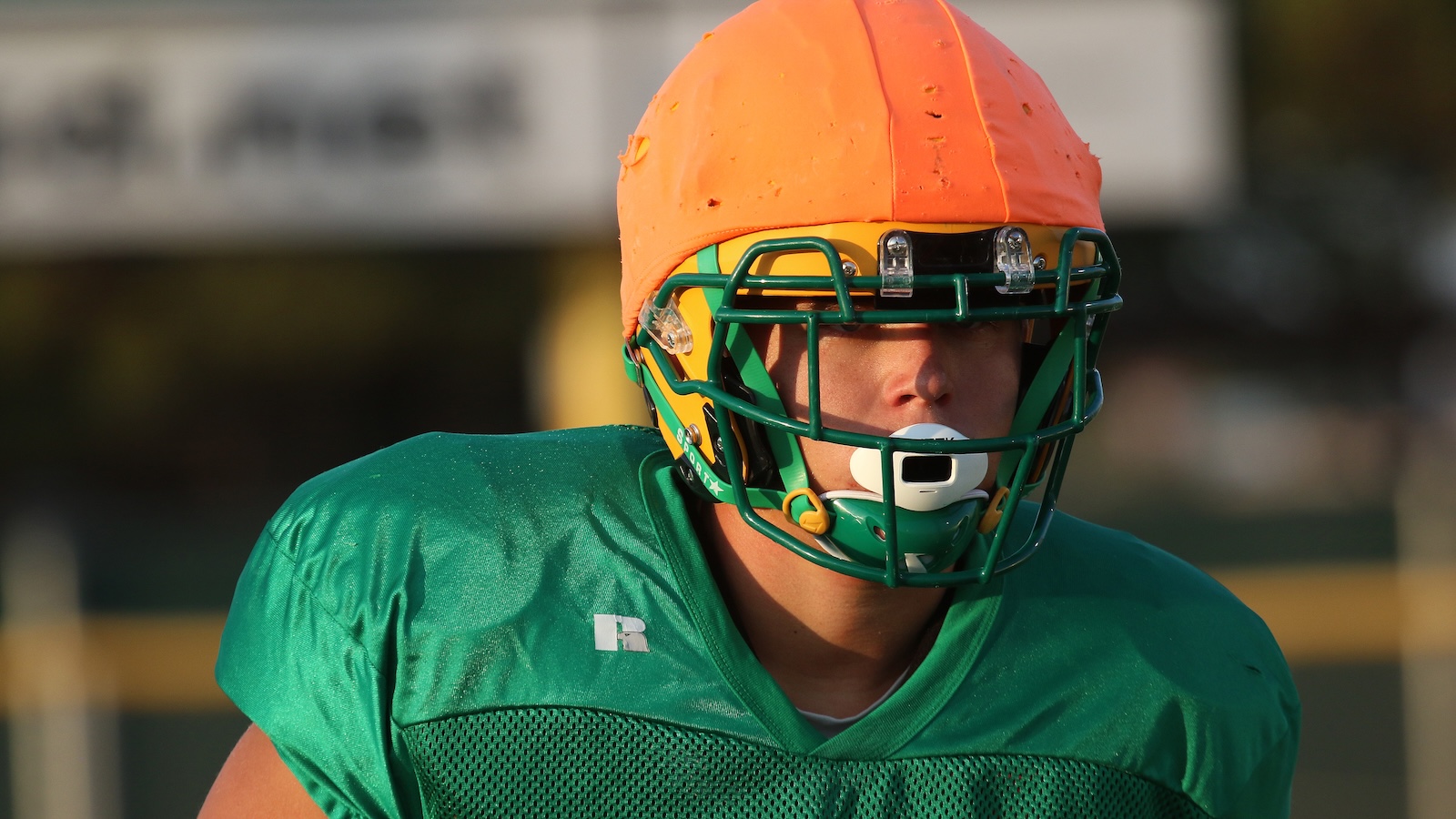 American football player in a green jersey wearing an orange helmet with a green face mask and white mouthguard during practice.