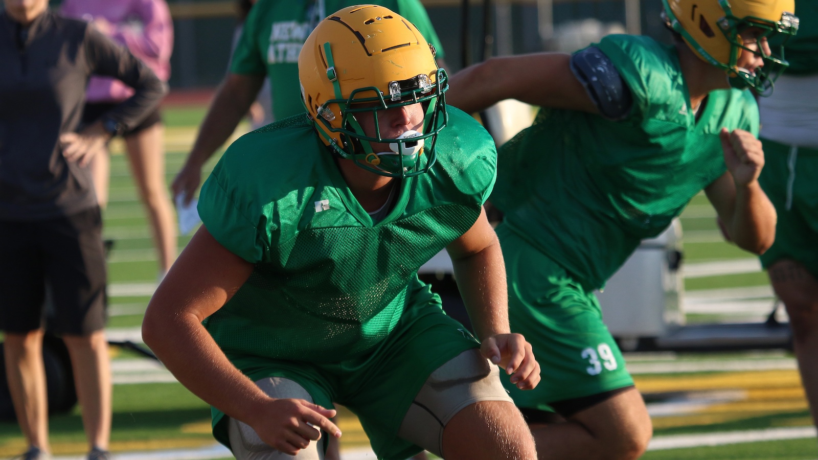 Football players in green jerseys practicing on a field, sprinting in formation with yellow helmets.