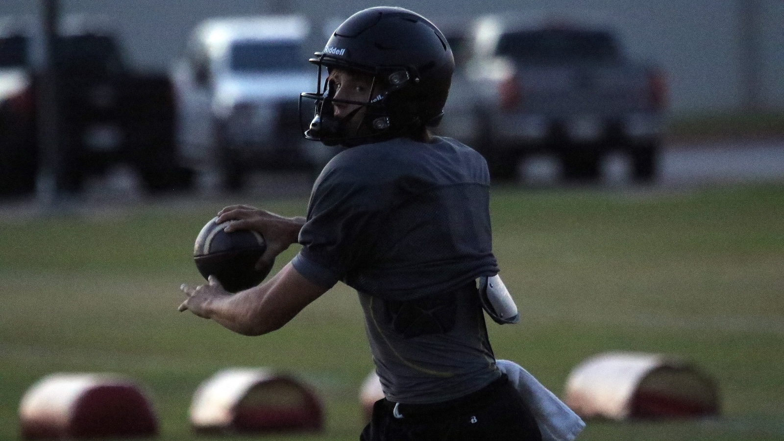 Quarterback in a dark helmet and gray jersey prepares to throw a football during a dusk practice, with practice cones in the foreground.