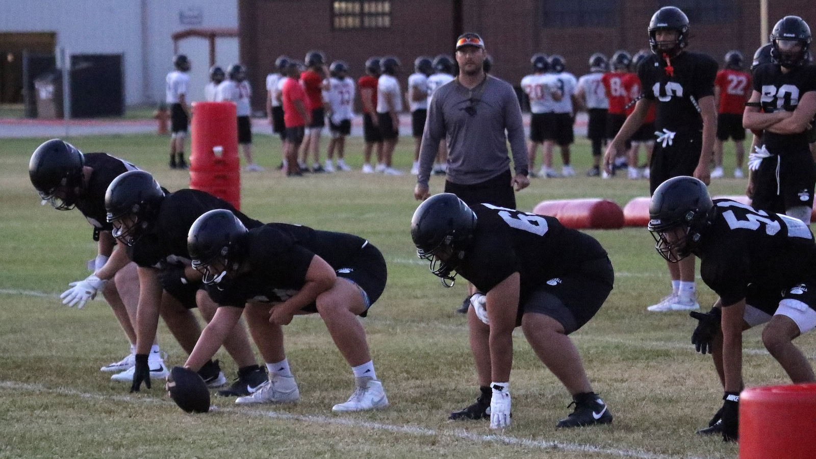 High school football offensive line in black uniforms crouches at the line, center about to snap, coach in gray shirt watching in the background.